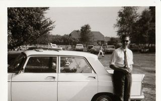 Een jonge man in een wit overhemd met opgerolde mouwen leunt lachend tegen een auto.