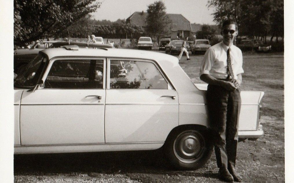 Een jonge man in een wit overhemd met opgerolde mouwen leunt lachend tegen een auto.