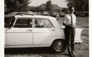 Een jonge man in een wit overhemd met opgerolde mouwen leunt lachend tegen een auto.