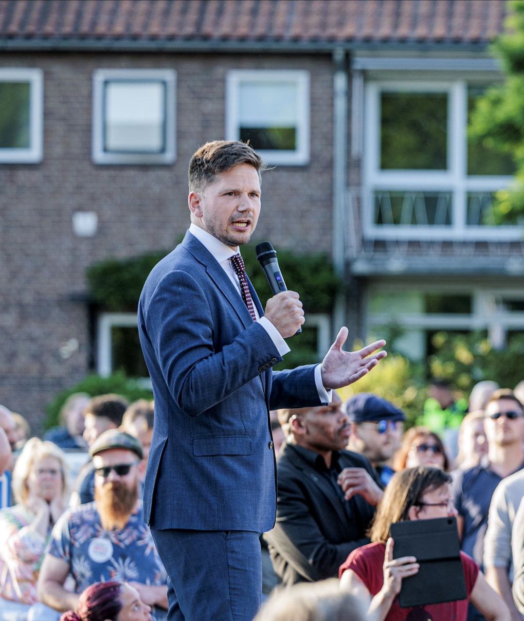 Man staat in de buitenlucht op een podium. Om hem heen staan mensen.