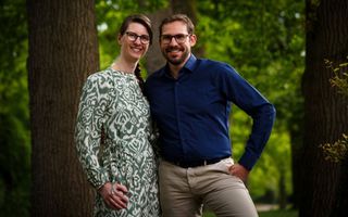 Een vrouw in een bewerkte jurk en een man in blouse staan samen tussen twee bomen, in een bos.