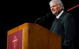 Een oude man met een zwart pak staat achter een houten katheder.