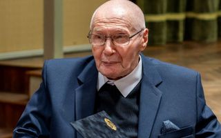 Een oude man met bril en gekleed in een blauw pak zit met een map op zijn schoot.