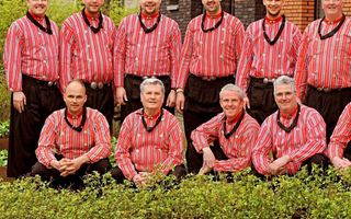 Groep mannen in zwarte broeken en rood-witte overhemden staan in twee rijen. De voorste rij zit op de hurken, de achterste rij staat.