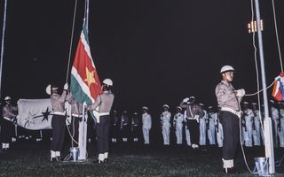 oude foto waarop zwarte mannen met witte helmen een surinaamse vlag hijsen. De vlag is groen met rode balk in het midden, in het rood staat een gele ster.