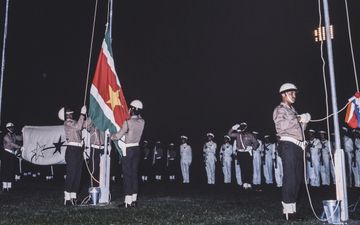 oude foto waarop zwarte mannen met witte helmen een surinaamse vlag hijsen. De vlag is groen met rode balk in het midden, in het rood staat een gele ster.