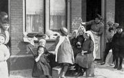 Woonomstandigheden, armoede: Kinderen spelen op de Baangracht in de Amsterdamse buurt De Jordaan. Een moeder kijkt vanuit de deuropening toe. Op de muur staan krijttekens. Nederland, datum onbekend [omstreeks 1910]. Origineel bevat een raster. Maker fotog