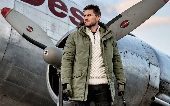 Man met winterjas poseert bij historisch vliegtuig.