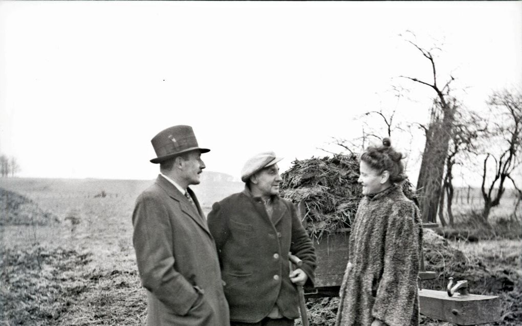 Zwart-witfoto van een goedgekleed echtpaar bij een boer die op het land bezig is om mest op een wagen te scheppen.