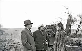 Zwart-witfoto van een goedgekleed echtpaar bij een boer die op het land bezig is om mest op een wagen te scheppen.