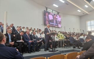 Man in grijs kostuum staat op podium en spreekt zaal mensen toe. Achter hem zit een koor in zwart-witte kleding.