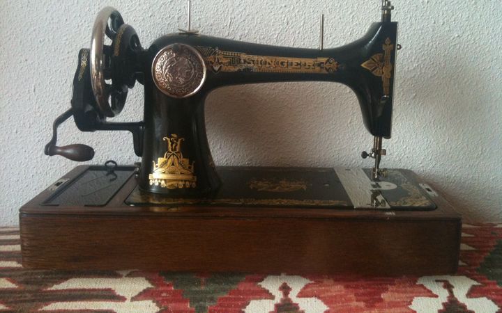 Foto van een ouderwetse naaimachine met links een slinger die met de hand wordt bediend. De naaimachine staat op een rood kleed met verschillende motieven.
