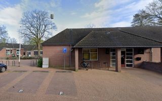 Een entree van een laag gebouw met bakstenen gevel en pannendak. Op de voorgrond parkeerplaatsen.