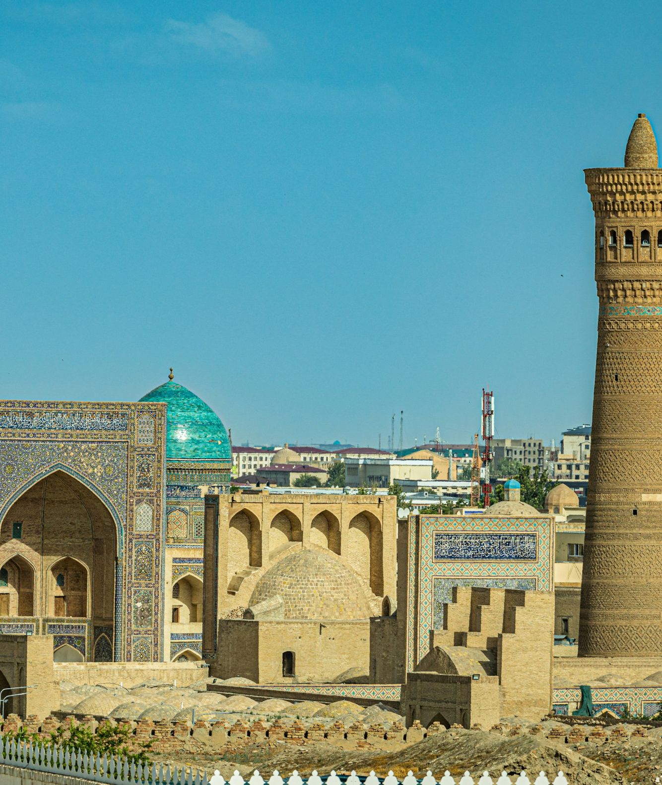 Een oud, bruin gebouwencomplex met turqoise koepels en een zandbruine minaret, tegen een felblauwe lucht. 