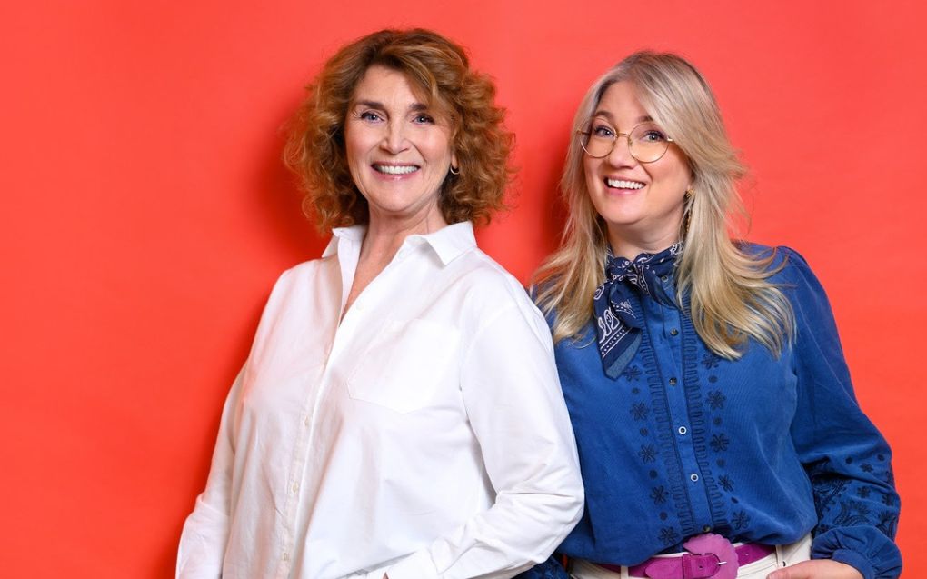 Twee vrouwen poseren tegen een oranjerode achtergrond. De dame links heeft kastanjebruine krullen tot op haar schouders en draagt een witte blouse. De dame rechts heeft lang, blond haar, een bril en draagt een jeansblauwe blouse met sjaaltje.