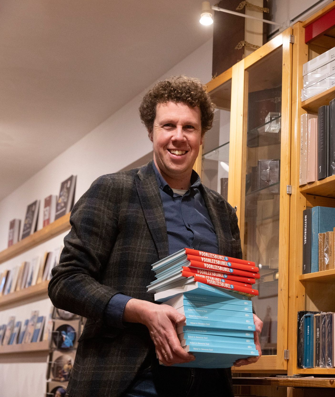 Een man met donker krullend haar houdt een stapel blauwe en rode boeken vast. Achter hem staat een lichte eiken kast vol boeken. 