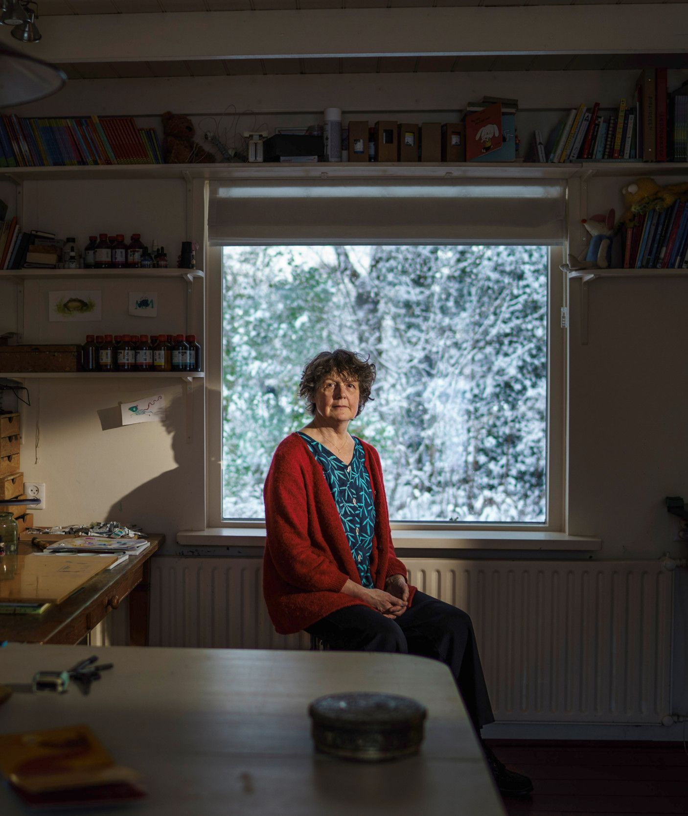 Donkere foto van een vrouw met kort, bruin haar en een rood vest aan. Ze zit op een kruk voor een raam met uitzicht op besneeuwde takken.  