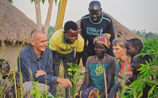 Nederlands boerenechtpaar en Congolese boeren en kinderen zitten in het gras. Op de achtergrond twee hutten.