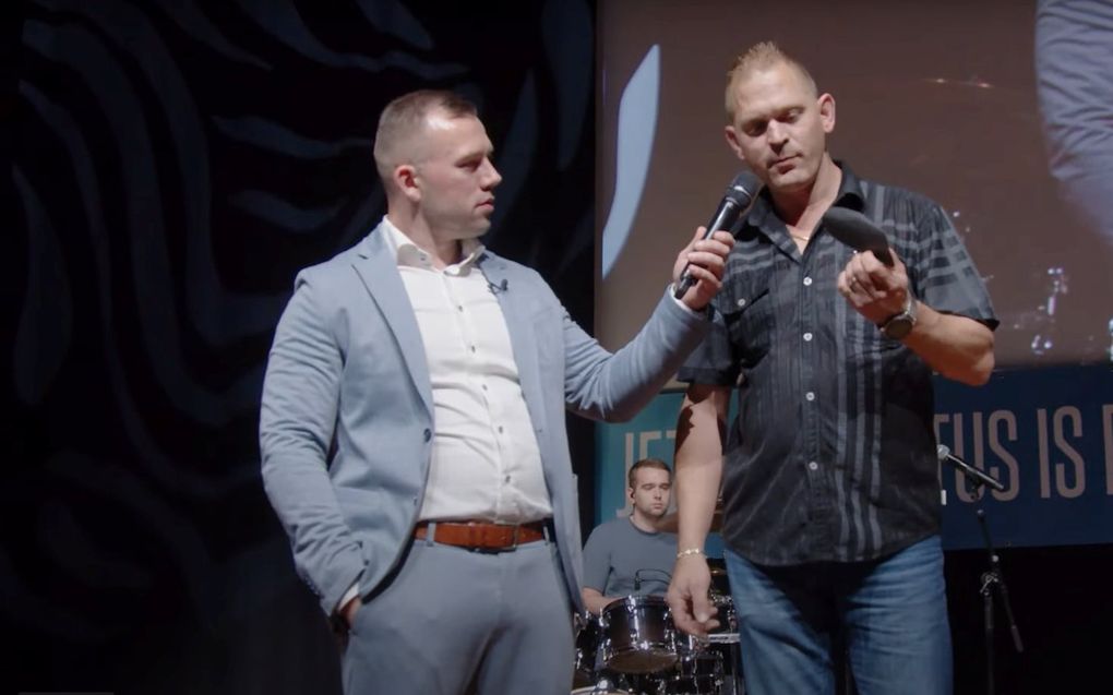 Man met microfoon in gesprek met een andere man. Op de achtergrond een man met gitaar en een drummer.