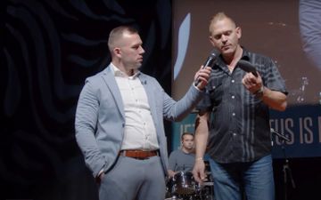 Voorganger Tom de Wal, in lichtblauw pak, houdt een microfoon voor een grote kerel in een ruitblouse die een steunzool omhooghoudt. 