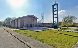 Vrij modern kerkgebouw met op de linkermuur een groot kruis. Rechts een vlag met daarop de naam Goede Herderkerk, en een open klokkentoren.