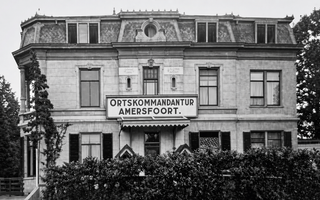 Zwart-wit foto van een stenen gebouw van drie verdiepingen met een heg ervoor. De ramen beneden hebben luiken. Boven de voordeur een bord met daarop: ”Ortskommandantur Amersfoort”.