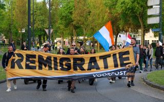 Een groep mensen loopt over straat met in hun hand een spandoek. Daarop staat met dikke letters ‘remigratie’. In de stoet worden ook Nederlandse vlaggen meegevoerd. Op de achtergrond wat publiek en politiemensen.