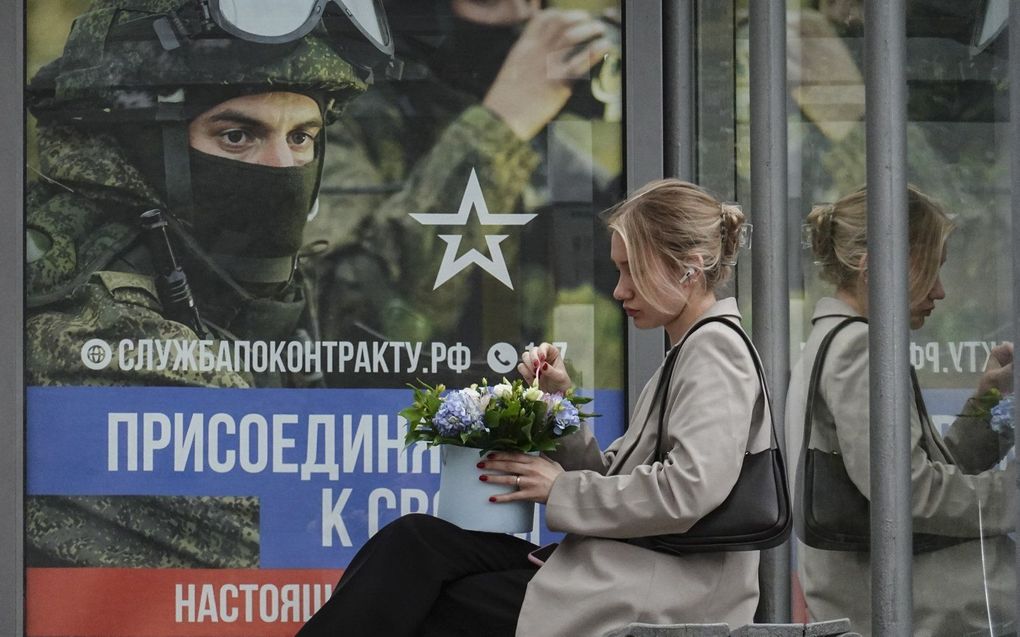 Een Russische vrouw zit in een bushalte voor een wervingsposter van het Russische leger. beeld EPA, YURI KOCHETKOV