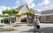 Koster Aart van den Dool en zijn vrouw Betsie voor het kerkgebouw van de christelijke gereformeerde kerk in Noordeloos. De kerk ligt buiten het dorp, aan de Noordzijde, parallel aan de rivier. Links naast de kerk bevindt zich de pastorie, rechts de koster