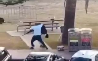 Man in wit shirt grijpt met zijn rechterhand om de nek van de in het zwart geklede schutter en pakt tegelijkertijd met zijn linkerhand een lang vuurwapen vast.