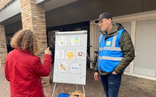 Een persoon met een rode jas aan kijkt naar een bord met een rebus. Daarnaast staat een man met een blauw hesje over zijn jas en een pet op.