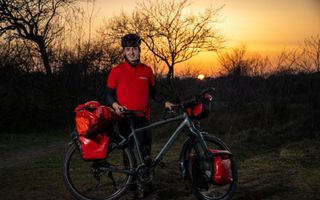 Een man met een rood T-shirt en een fietshelm staat met zijn fiets aan de hand buiten, voor een ondergaande zon.