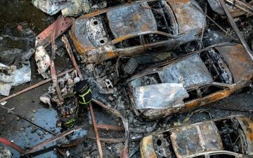 Drie verbrande auto's met daarbij mannen in zwarte pakken met gele strepen, vanboven gefotografeerd.