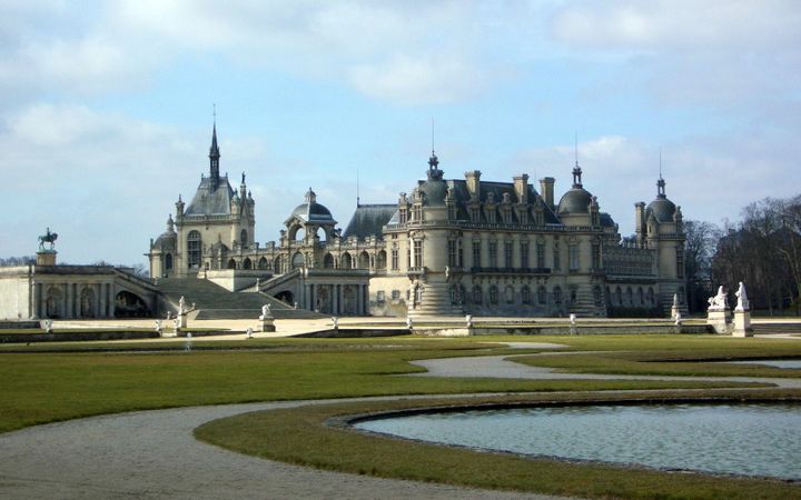 Kasteel van wit gesteente met leien daken. Het kasteel heeft verschillende torentjes en zuilen. Op de voorgrond is een vijver te zien met enkele standbeelden.