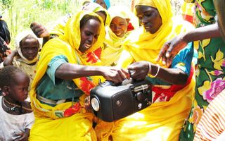 Vrouwen en kinderen zitten rondom een radio, die twee vrouwen in gele kleding vasthouden. 