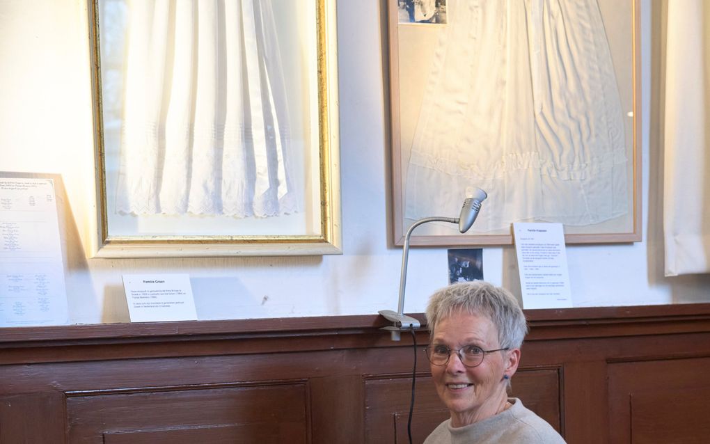 Een oudere vrouw, bril, lachend, bij een doopjurk.