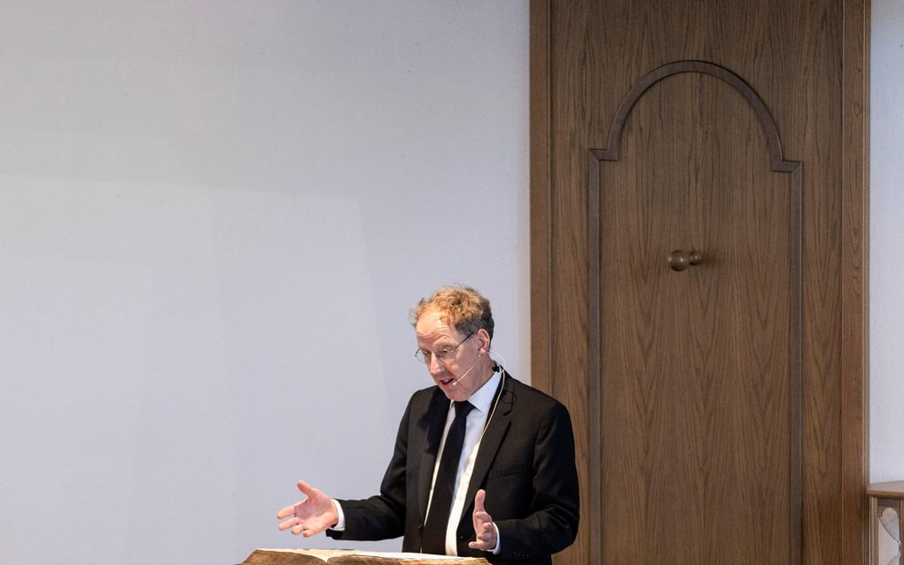Een man met donkerblond haar, bril en zwart pak spreekt op een kansel.
