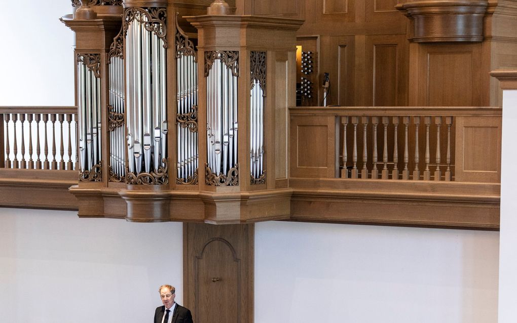 Een man met donkerblond haar, bril en zwart pak spreekt op een kansel.