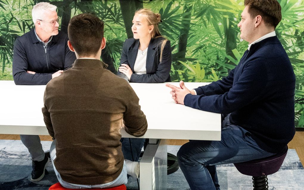 Een aantal mensen zitten rondom een tafel. Ze zitten op ergonomische stoelen, met een aparte vorm en veren.