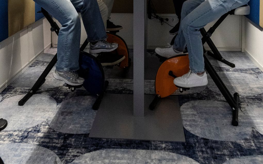 Drie mannen en een vrouw zitten aan een witte tafel. Aan de kop van de tafel hangt een zwart scherm. Het viertal zit in een ruimte van houten schermen met blauwe vlakken.