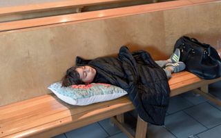 Een kind ligt met een kussen onder een jas te slapen op een houten kerkbank tegen een lage afscheiding van hout. Op de achtergrond een witte muur met een nis met bakstenen rondboog en daarin een groot houten kruis, een knielbank en twee stoelen.