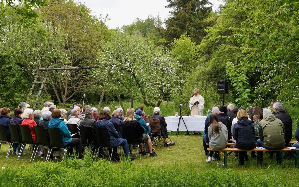Op een grasveld staan rijen stoelen en banken met mensen erop. Vooraan staat een man in een lang wit gewaad die uit de Bijbel leest.