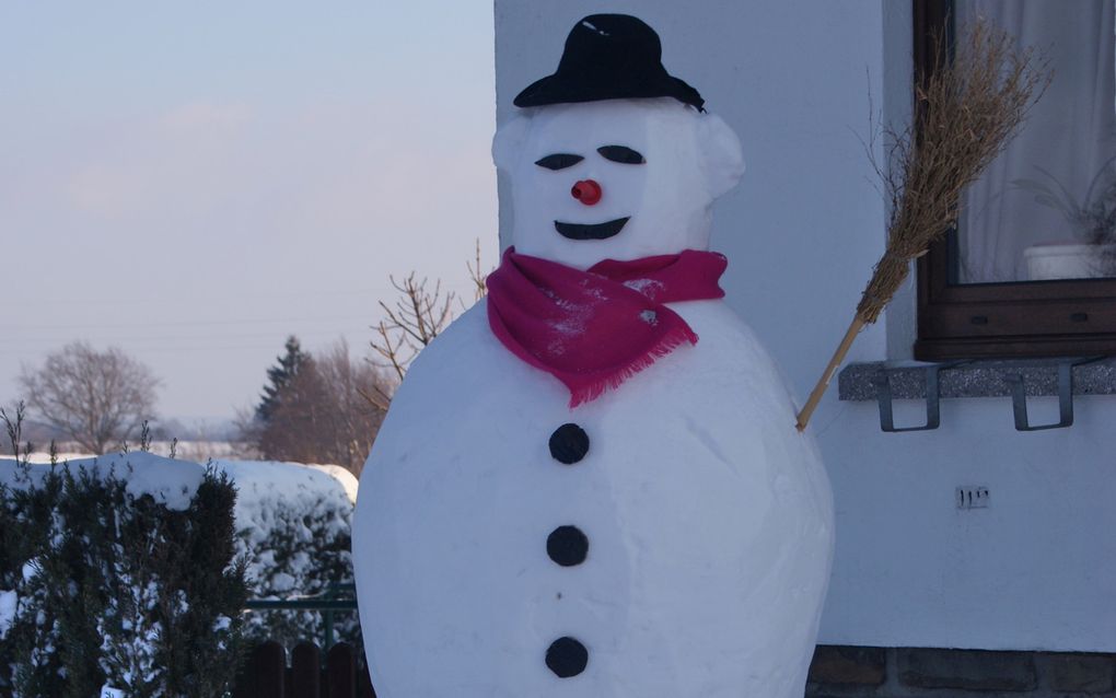 Sneeuwpop tegen een wit huis. Rode sjaal, hoedje, grote zwarte knopen.