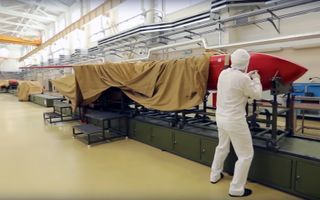 Een man in een wit pak staat in een loods bij een rode raket, die deels met een bruine doek is afgedekt 