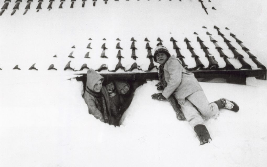 Zwart-witfoto van drie kinderen onder een besneeuwde dakrand in een gat in de sneeuw. Een kind zit ernaast op een sneeuwhoop.