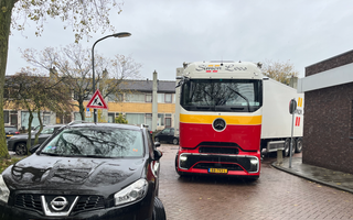 Vrachtwagen met veelkleurige voorkant staat vast in een bocht vanwege fout geparkeerde auto’s, waaronder een zwarte Nissan.