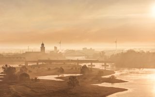 Uitzicht op een de stad Deventer in de ochtendzon. Op de voorgrond een rivier, in de verte in silhouet twee bruggen windmolens en twee kerken.