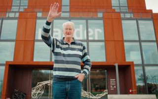 Oudere man in een gestreept shirt en spijkerbroek, staat zwaaiend voor een kantoorgebouw.