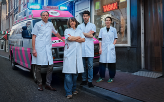 Artsen in witte jassen staan voor een roze ambulance in een winkelstraat.