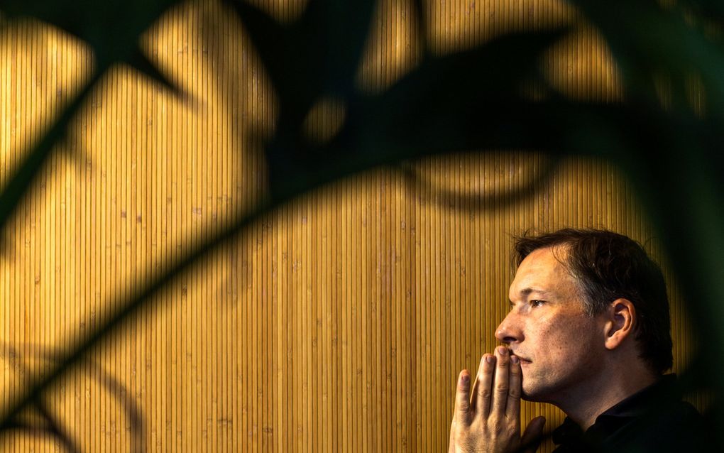 Man van middelbare leeftijd met donkerblauw overhemd heeft zijn handen tegen elkaar gedrukt tegen zijn kin aan. De man staart in de verte; achter hem is een houtkleurige wand